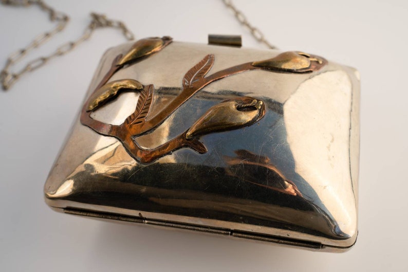 May include: A silver metal clutch bag with a chain strap. The bag features a floral design with copper and brass accents.