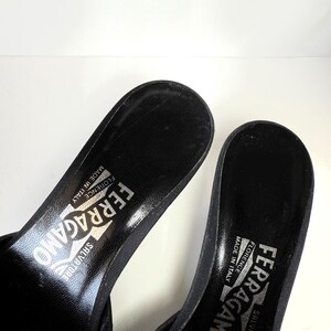 May include: Close-up of two black Salvatore Ferragamo sandals. The sandals have a textured insole and the brand name is printed in silver. The sandals are made in Florence, Italy.