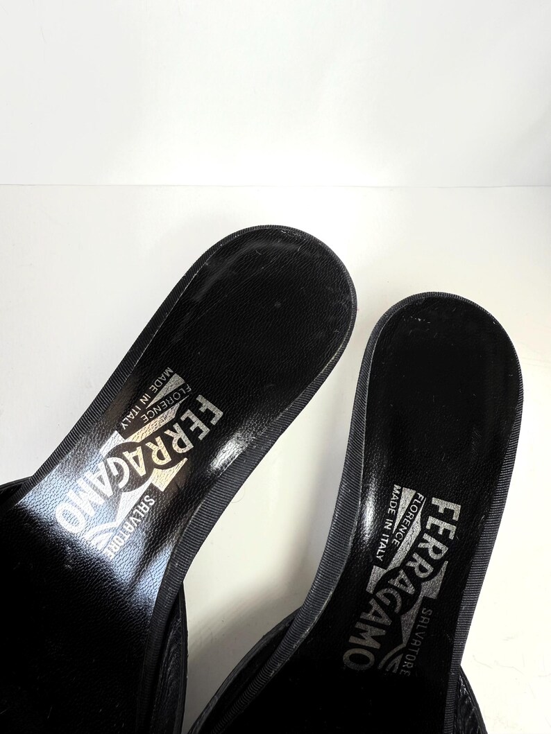 May include: Close-up of two black Salvatore Ferragamo sandals. The sandals have a textured insole and the brand name is printed in silver. The sandals are made in Florence, Italy.