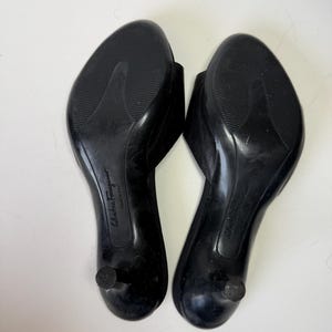 May include: A pair of black leather heeled sandals, viewed from the soles. The sandals have a rounded toe and a small, rounded heel. The soles are textured for grip. The brand name "Salvatore Ferragamo" is visible.