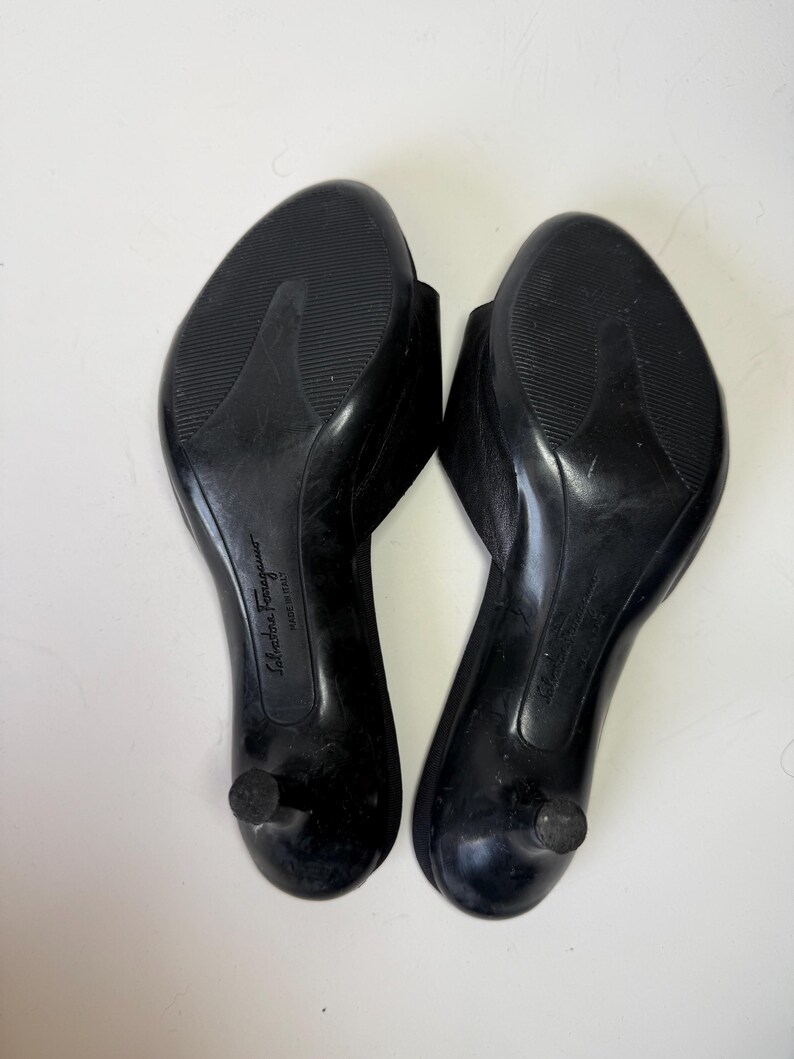May include: A pair of black leather heeled sandals, viewed from the soles. The sandals have a rounded toe and a small, rounded heel. The soles are textured for grip. The brand name "Salvatore Ferragamo" is visible.