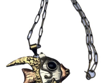 Könnte beinhalten: Eine Halskette mit einem Fisch-Anhänger aus Gold, Silber und Kupfer mit einem schwarzen Steinauge. Die Kette ist aus Goldmetall.