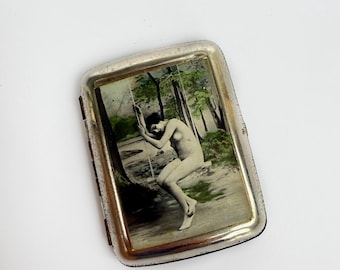 Pode incluir: Estojo de cigarro vintage em tom prateado com formato retangular. O estojo apresenta uma imagem pintada de uma figura nua balançando perto da água, cercada por árvores. O estojo tem uma tampa articulada e um interior preto.