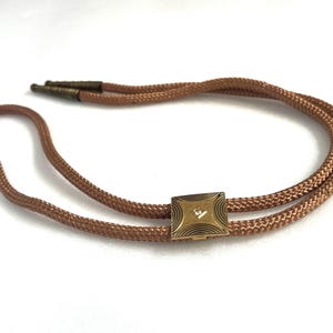 May include: A vintage bolo tie with a woven brown cord. The tie features a square, gold-toned slide with a diamond-shaped accent in the center. The slide has a black, geometric design around the edges.
