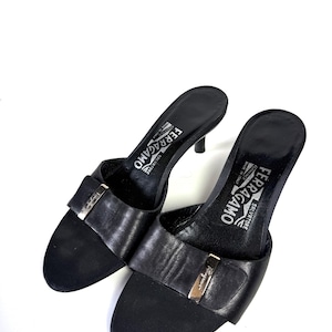 May include: A pair of black Salvatore Ferragamo heeled sandals. The sandals feature a leather strap with a silver-tone buckle and a small heel. The brand name is visible on the insole. The shoes are set against a white background.