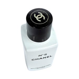 May include: A white rectangular bottle of Chanel No. 5 body lotion with a black cap. The cap features a white interlocking double-C logo. The product name and brand are printed in black on the front of the bottle.