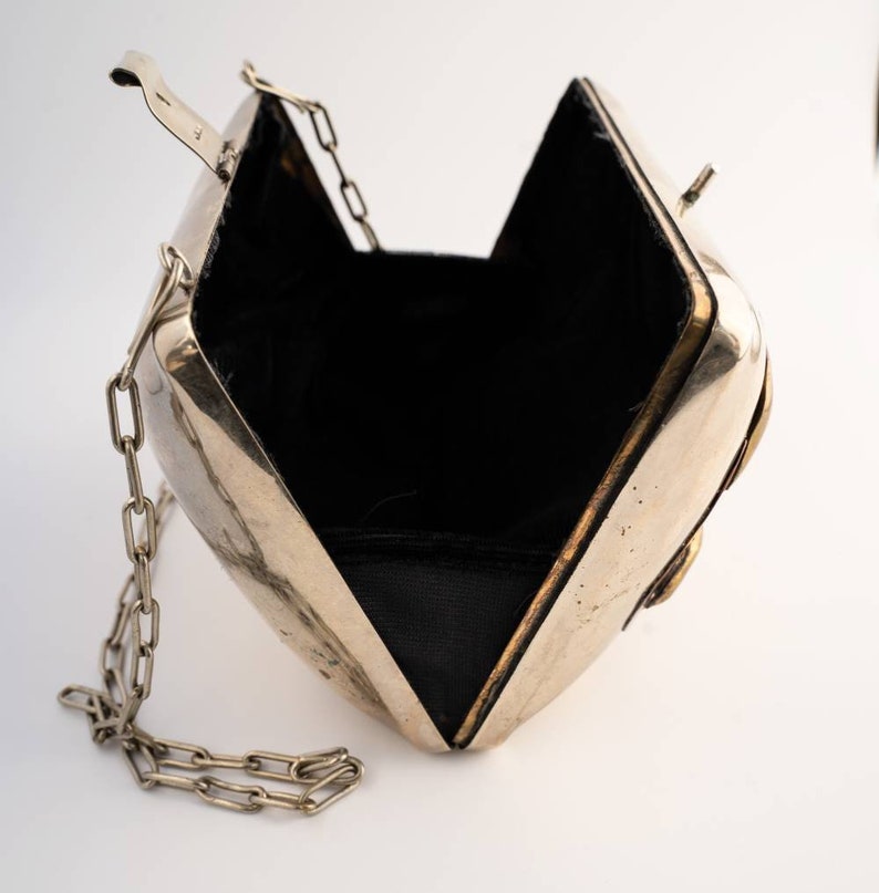 May include: A silver metal clutch bag with a chain strap and a black velvet lining. The bag is open and the lining is visible.