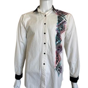May include: White button-down shirt with long sleeves, black collar, cuffs, and buttons. A vertical design on the left side features geometric patterns in black, teal, purple, and pink. The shirt is displayed on a mannequin.