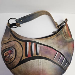 Könnte beinhalten: Eine braune Ledertasche im Hobo-Stil mit einem farbenfrohen, abstrakten Design, das auf die Vorderseite gemalt ist. Die Tasche hat einen langen Riemen und einen Reißverschluss.