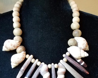 Puede incluir: Un collar de cuentas de madera marrón con una variedad de conchas marinas, incluyendo conchas blancas, marrones y moradas. El collar se muestra en un maniquí negro.