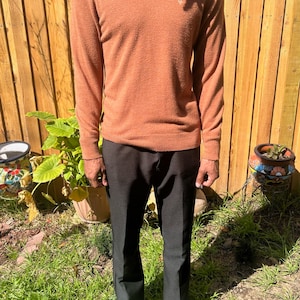 Può includere: Una persona indossa un maglione a maniche lunghe color ruggine con una collana d'argento e pantaloni neri. Indossa scarpe da ginnastica nere e si trova davanti a una recinzione di legno.