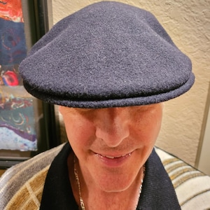 May include: A dark blue wool newsboy cap with a brim. The cap is worn by a person with a light complexion and a smile.