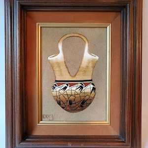 以下が含まれることがあります： ユニークなダブルスパウトデザインのベージュ色の花瓶が特徴の額装アートワーク。花瓶は、黒、赤、ベージュの幾何学模様で飾られています。アートワークはニュートラルな背景に配置され、濃い茶色の木製フレームに額装されています。