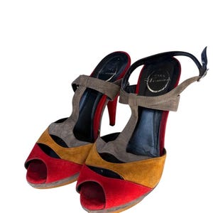 Può includere: Sandali con tacco alto aperti in punta con un design a blocchi di colore. I sandali presentano pannelli in pelle scamosciata rossa, giallo senape e grigia. I tacchi sono rossi e i cinturini grigi. Il marchio "ZARA Woman" è visibile sulla soletta.