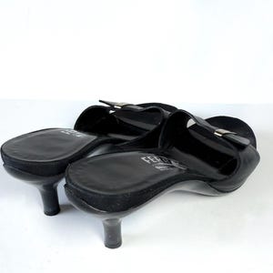 May include: Black leather slip-on heels with a small heel. The shoes have a rounded toe and a strap across the top with a silver-colored buckle. The heels are approximately 2 inches tall.