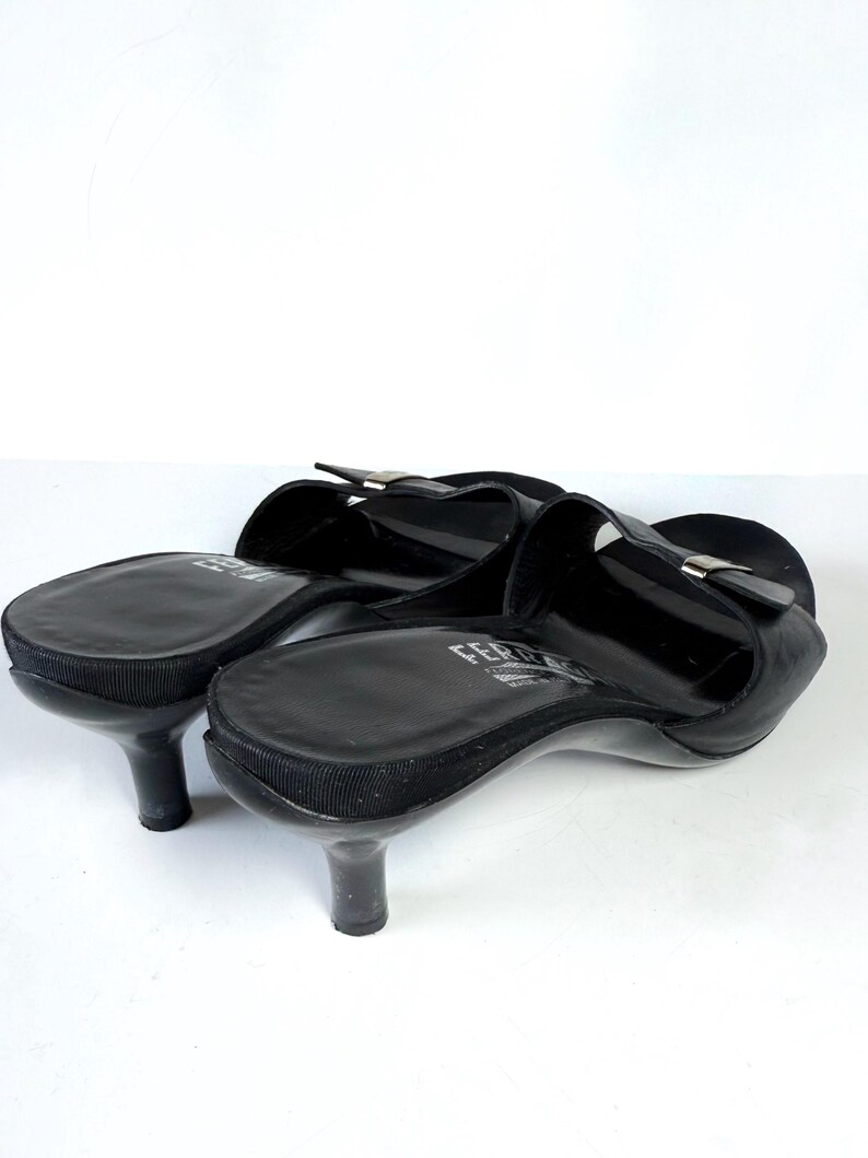 May include: Black leather slip-on heels with a small heel. The shoes have a rounded toe and a strap across the top with a silver-colored buckle. The heels are approximately 2 inches tall.