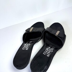 May include: A pair of black leather Salvatore Ferragamo slides. Each slide features a silver-tone buckle and the brand name printed in white. The sandals are set against a white background.