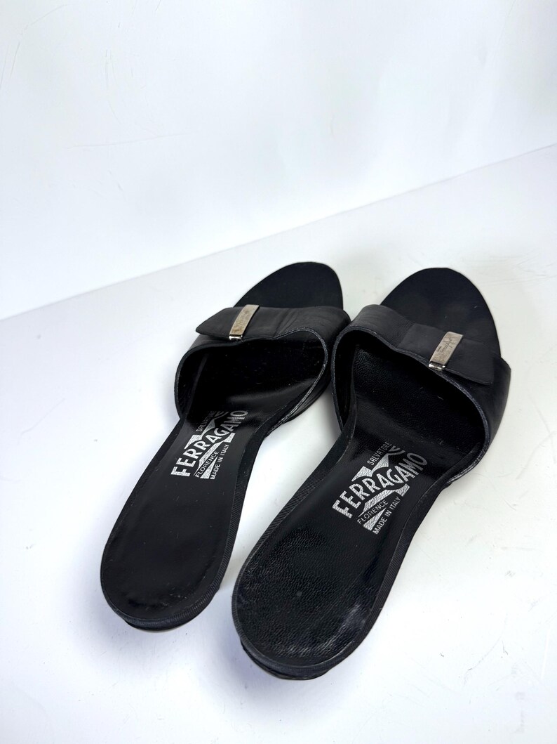 May include: A pair of black leather Salvatore Ferragamo slides. Each slide features a silver-tone buckle and the brand name printed in white. The sandals are set against a white background.