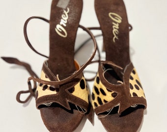 May include: A pair of brown suede sandals with a cheetah print design on the front straps. The sandals have a high heel and ankle straps. The word "Orex" is printed on the insole of each shoe.