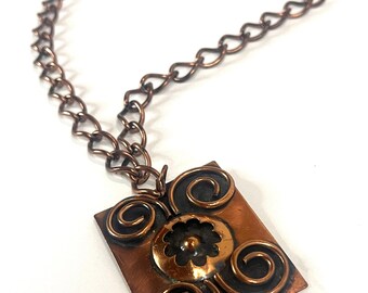 May include: A copper-toned necklace with a square pendant. The pendant features a central floral design and swirling accents. The chain is made of interlocking links. The necklace is displayed against a white background.