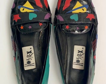 May include: A pair of black leather loafers with a pointed toe. The shoes have a colorful design with stars, hearts, and other geometric shapes. The brand name "Astrx" is printed on the inside of each shoe.