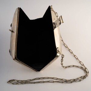 May include: A silver metal clutch bag with a chain strap. The bag is open and the inside is lined with black fabric.
