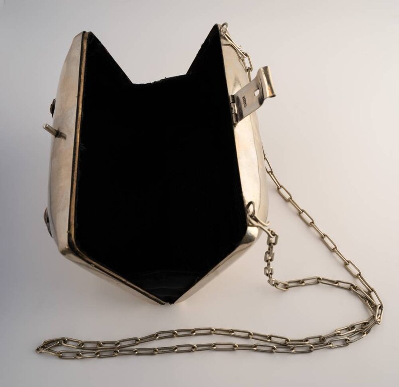 May include: A silver metal clutch bag with a chain strap. The bag is open and the inside is lined with black fabric.