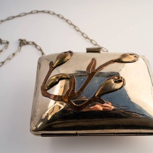 May include: A silver clutch bag with a chain strap and a floral design made of copper and brass.