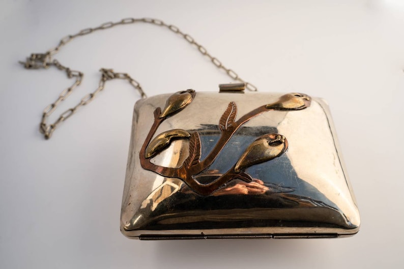 May include: A silver clutch bag with a chain strap and a floral design made of copper and brass.