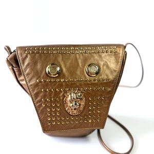 May include: A metallic gold-colored crossbody bag with a triangular shape. The bag features gold-colored studs and two circular gold-colored buttons. A gold-colored lion's head is centered on the front.