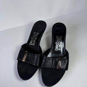 May include: A pair of black leather Salvatore Ferragamo heeled sandals. The sandals have a rounded toe and a strap across the top with a gold-colored buckle. The heels are approximately 3 inches high. The brand name is visible on the insole.