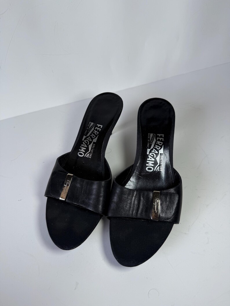 May include: A pair of black leather Salvatore Ferragamo heeled sandals. The sandals have a rounded toe and a strap across the top with a gold-colored buckle. The heels are approximately 3 inches high. The brand name is visible on the insole.