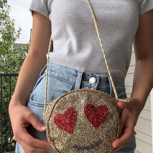 Puede incluir: Un bolso bandolera redondo con purpurina dorada con ojos en forma de corazón rojos y una sonrisa cosida. El bolso tiene una fina correa de cuerda multicolor y un cierre de cremallera. El bolso tiene unos 18 cm de diámetro.