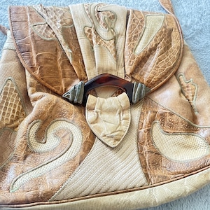 May include: A brown leather handbag with a unique patchwork design. The bag has a flap closure with a decorative brown and gold clasp. The bag is made of different types of leather, including crocodile embossed leather.