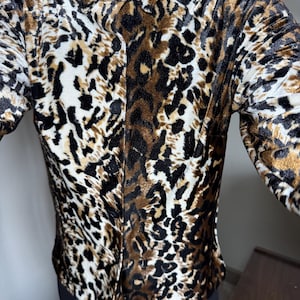 Puede incluir: Camisa con estampado de leopardo en tonos marrón, negro y crema. La camisa de manga larga tiene cuello y botones negros. El estampado animal se ve en la tela.