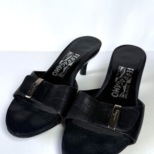 May include: A pair of black leather Salvatore Ferragamo heeled sandals. Each sandal features a strap with a silver-tone buckle and a small heel. The brand name is visible on the insole.