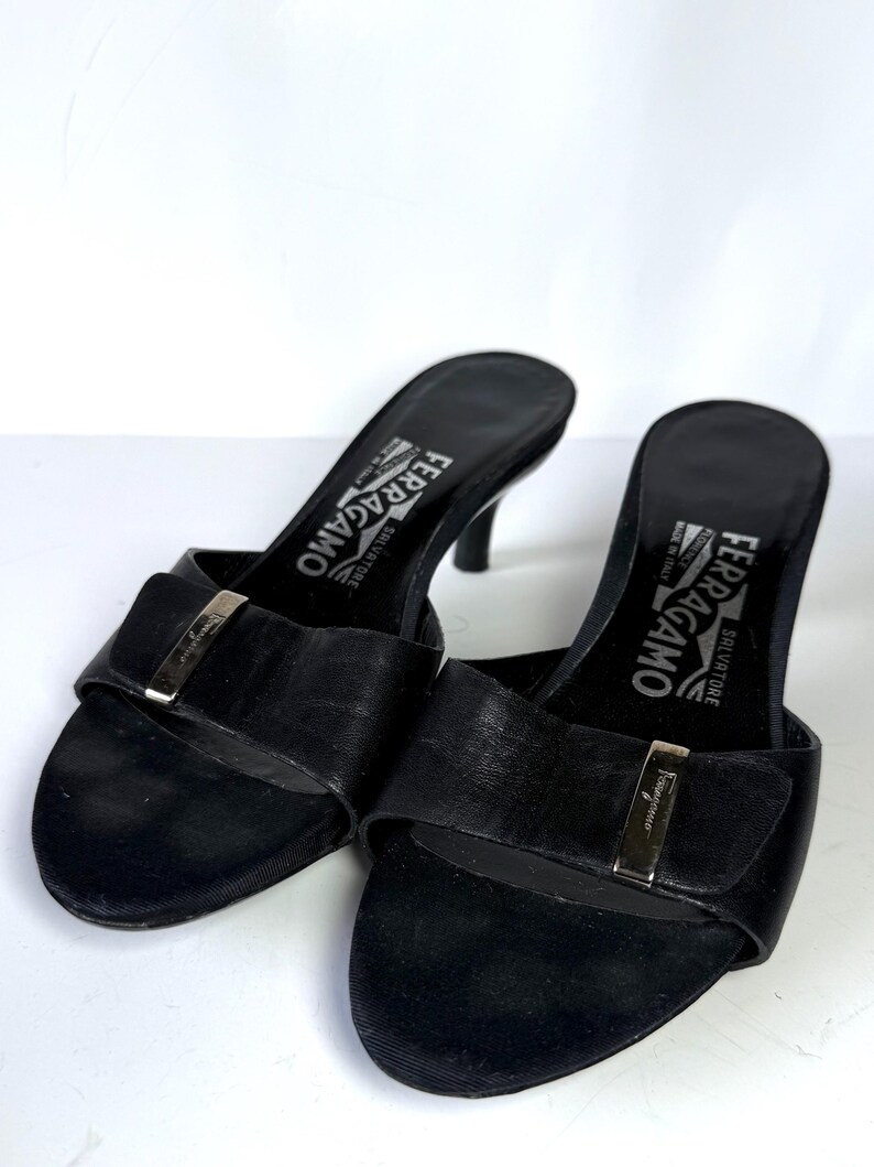 May include: A pair of black leather Salvatore Ferragamo heeled sandals. Each sandal features a strap with a silver-tone buckle and a small heel. The brand name is visible on the insole.
