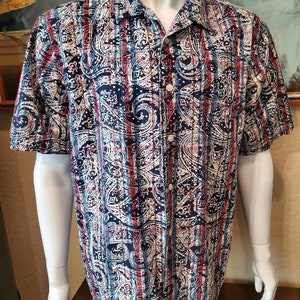 Pode incluir: Uma camisa de manga curta com botões e um padrão paisley e listrado em tons de azul, vermelho e branco.
