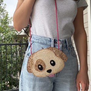 Puede incluir: Un bolso cruzado con forma de perro y correa rosa. El bolso es beige con detalles brillantes, ojos negros y una lengua rosa. La correa es un cordón rosa brillante.
