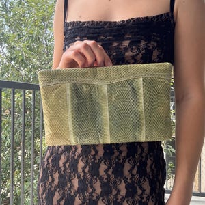 May include: A light green clutch with a snakeskin pattern. The rectangular bag has a segmented design and a neutral color palette. The clutch is held in front of a black lace dress.