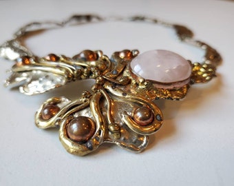 Puede incluir: Un collar de plata, cobre y latón con una gran piedra de cuarzo rosa en el centro. El collar está intrincadamente diseñado con un motivo floral.