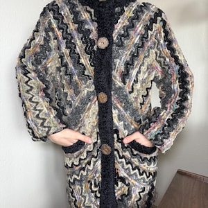 Op de afbeelding: Een lange, veelkleurige jas met een zigzagpatroon in grijstinten, zwart en beige. De jas heeft een zwarte kraag, bies en zakken, met grote houten knopen. De jas is ongeveer 102 cm lang.