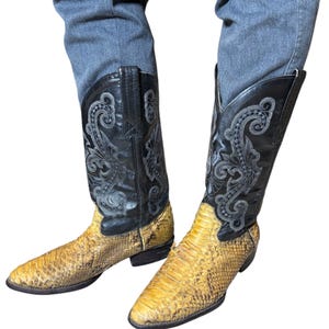 May include: A pair of cowboy boots with a snakeskin pattern on the foot and a black leather upper. The boots have decorative stitching and a pointed toe. The boots are worn with blue jeans.