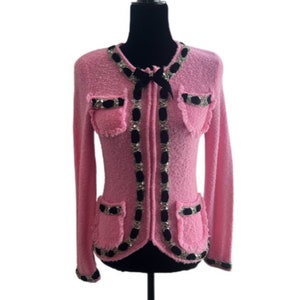 May include: A pink knitted cardigan with black trim and silver embellishments. The cardigan has a black velvet bow at the neckline and pockets on the front.