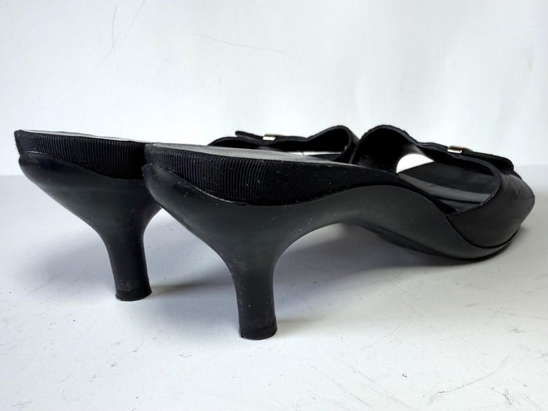 May include: Black open-toe heeled sandals with a bow detail. The sandals have a small heel and a textured fabric strap. The shoes are made of a shiny material and are set against a white background.