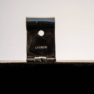 May include: Close-up of a silver metal hinge with a small hole and the word "LAMBERTS" engraved on it.
