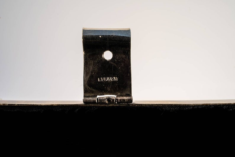 May include: Close-up of a silver metal hinge with a small hole and the word "LAMBERTS" engraved on it.
