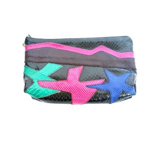 Peut inclure: Pochette noire à texture peau de serpent, ornée d'une bande rose en zigzag et d'étoiles turquoise, roses et bleues. La pochette a une fermeture éclair dorée et une forme rectangulaire.