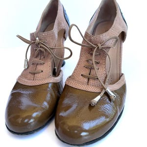 May include: A pair of two-tone women's shoes with a lace-up design. The shoes feature a glossy brown toe and heel, with a textured tan upper and a black accent at the heel. The shoes have a high heel and are set against a white background.