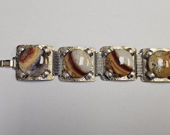 Pode incluir: Uma pulseira vintage com quatro elos quadrados dourados, cada um com uma pedra redonda polida. As pedras exibem uma mistura de cores marrom, bege e creme com padrões únicos. A pulseira tem um fecho.